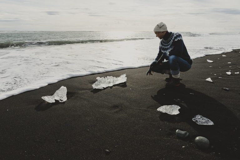 Person in warmer clothing beugt sich über Eisstücke am schwarzen Sandstrand.