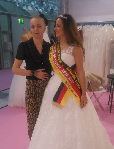 Eine Frau in Hochzeitskleid mit einer Miss-Wahl-Schärpe steht neben einer anderen Frau.