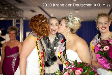 Drei Frauen mit Blumensträußen küssen sich freudig, im Hintergrund stehen weitere Teilnehmerinnen.